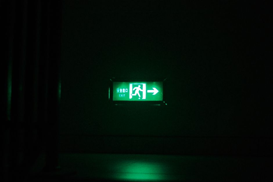 A green illuminated emergency exit sign is mounted on a dark wall indoors, indicating the direction to the nearest exit with an arrow pointing to the right. The sign features a running figure and char